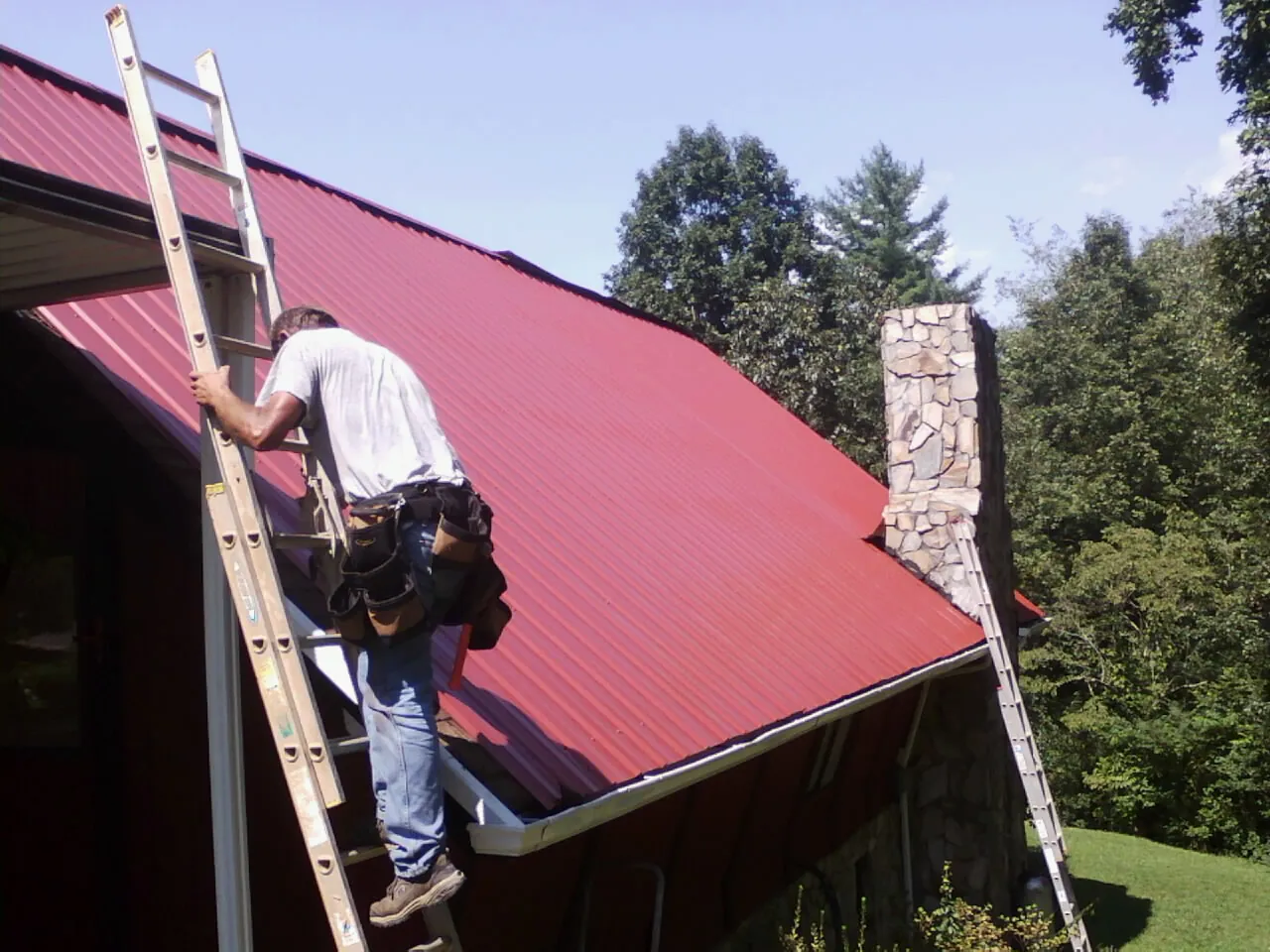Roofer on a ladder beside a red metal roof and stone chimney — Soffit & Fascia Repair by Roofing Brothers in Nezperce